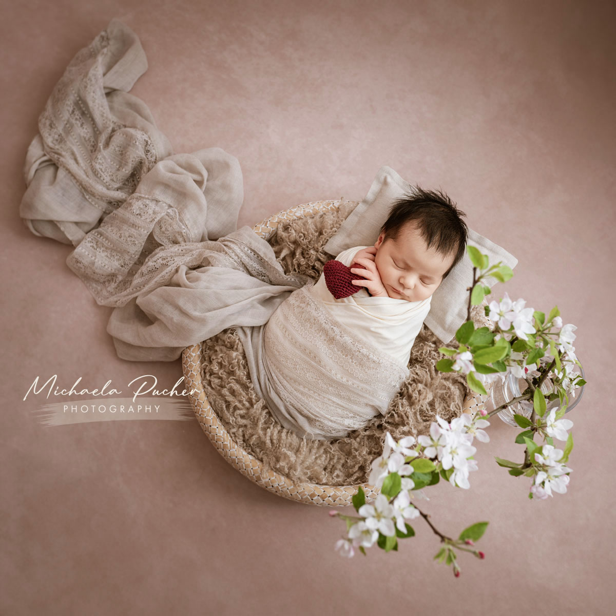 Neugeborenes eingewickelt in helle Tücher, liegend in einem weichen Körbchen, mit einem kleinen roten Häkelherz in der Hand und umrahmt von blühenden Zweigen.
